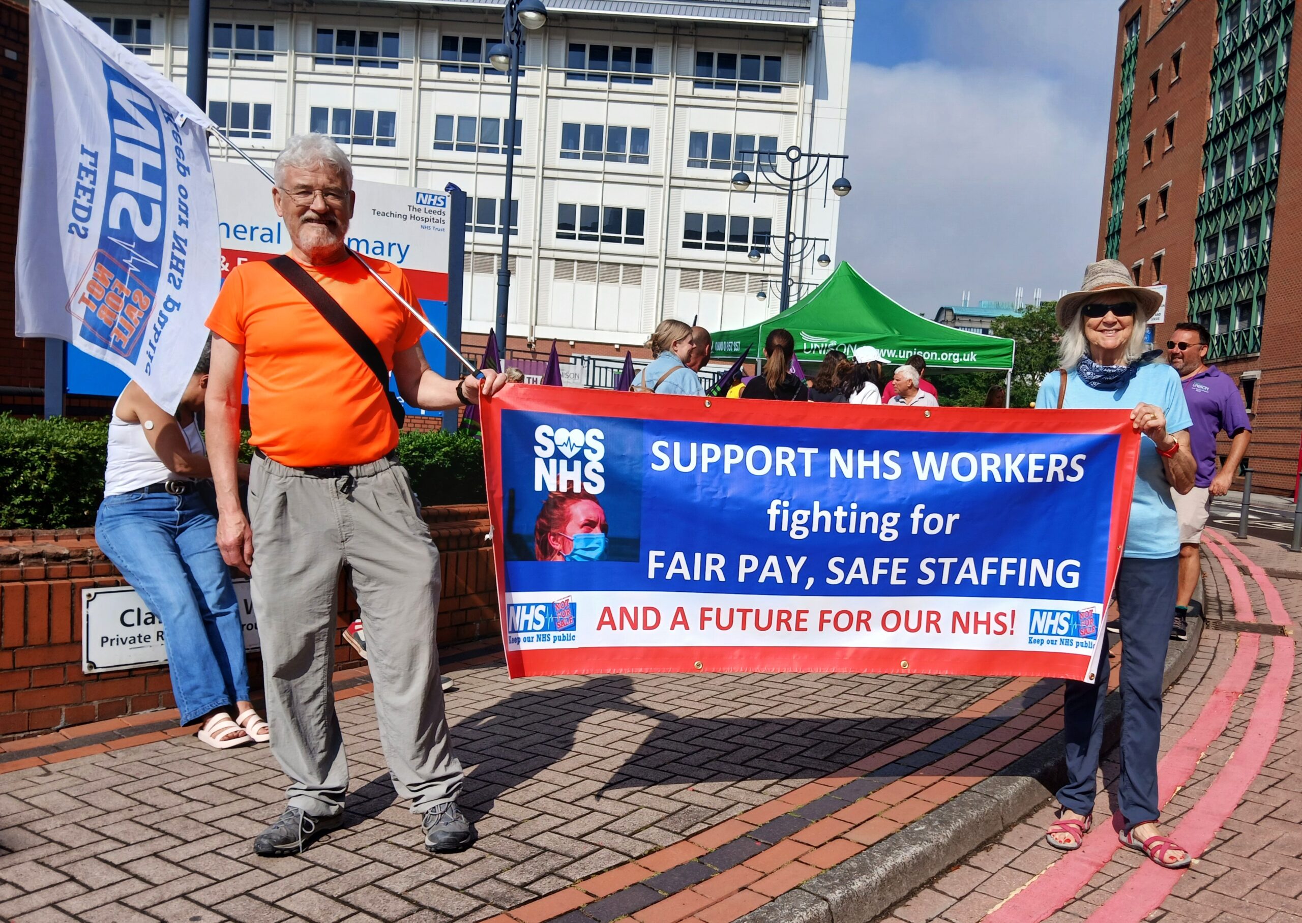 Two people outside holding a banner that states: Support NHS workers fighting for fair pay, safe staffing and a future for our NHS! SOS NHS and Keep Our NHS Public logos.