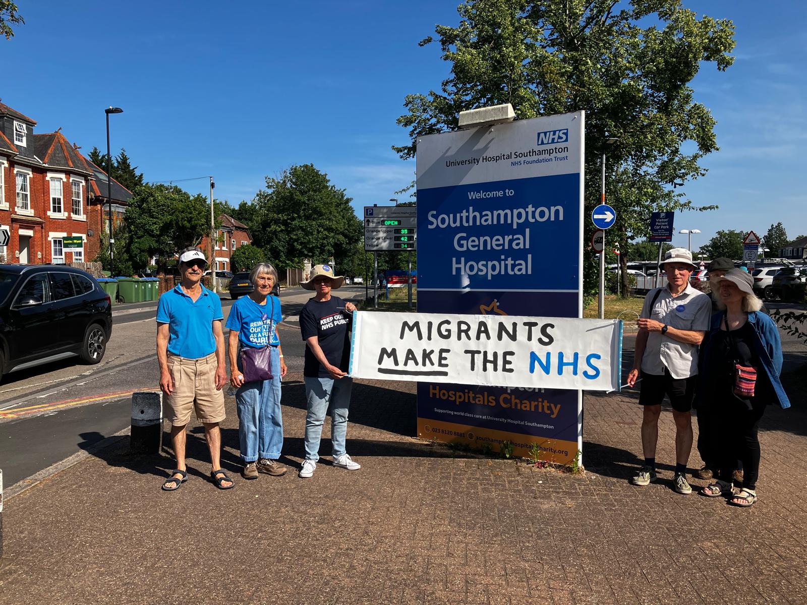 Four people pose in front of Southampton Hospital holding a banner that reads 'Migrants Make the NHS'